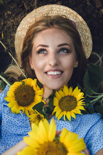 Long-haired blonde young woman in a straw hat and blue dress on a field of sunflowers. - Foto, afbeelding