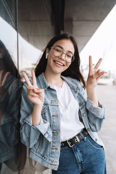 Happy young pretty woman student wearing eyeglasses walking outdoors resting listening music with earphones showing peace gesture. - 写真・画像