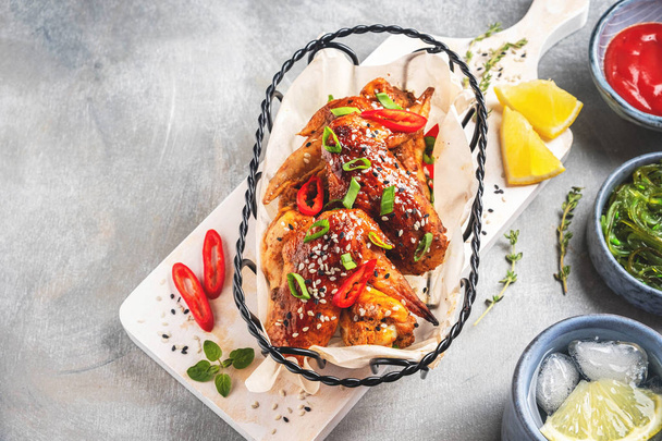 Grilled chicken wings in a metal basket on a white wooden board, seaweed, tomato sauce and mug of water with ice and lemon. Gray background, top view, copy space. - Foto, Imagem