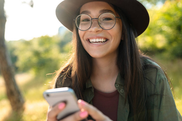 Photo of attractive laughing woman wearing stylish hat and eyeglasses using cellphone while walking in green park - Foto, afbeelding