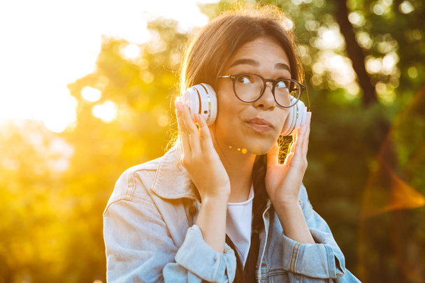 Thinking young teenage girl student sitting outdoors in beautiful green park listening music with headphones. - 写真・画像