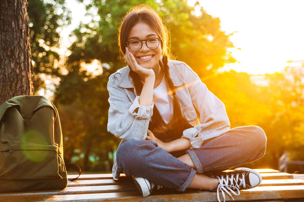 Happy cute young student girl wearing eyeglasses sitting on bench outdoors in nature park with beautiful sunlight. - Fotoğraf, Görsel