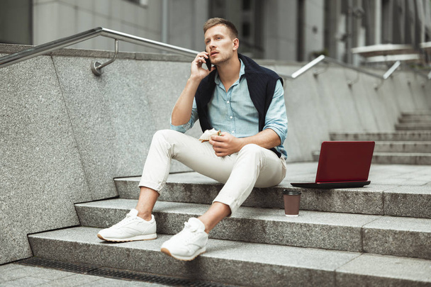 young handsome office worker speaking on the phone while eating his sandwich and drinking coffee for lunch during break sitting on the stairs with laptop - Foto, Bild
