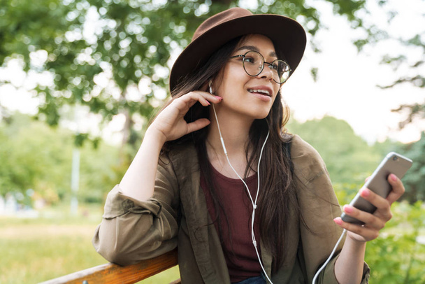 Photo of nice woman wearing hat and eyeglasses using earphones and smartphone in green park - Foto, Imagen