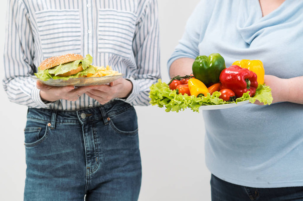 zwei Mädchen, dick und dünn, mit gesundem Essen aus Gemüse und Obst und ungesundem Fast Food mit einem Hamburger. das Konzept der Ernährung. - Foto, Bild