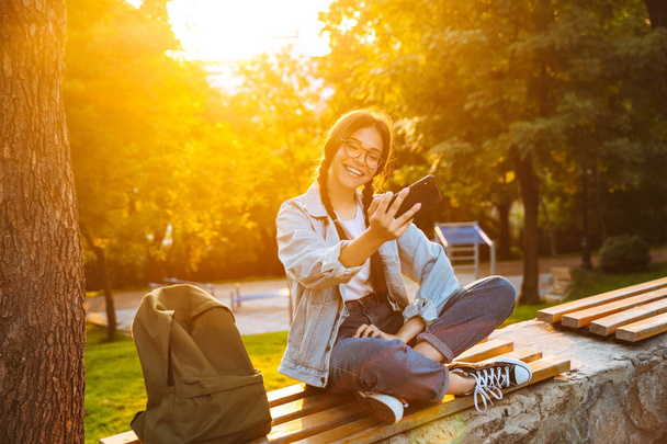 Souriant rire mignon jeune fille étudiante portant des lunettes de vue assis sur le banc à l'extérieur dans le parc naturel avec une belle lumière du soleil en utilisant téléphone mobile parler
. - Photo, image
