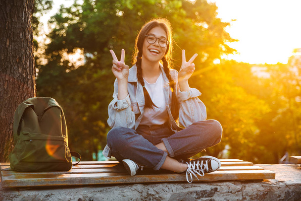 Cheerful cute young student girl wearing eyeglasses sitting on bench outdoors in nature park with beautiful sunlight showing peace gesture. - Photo, Image