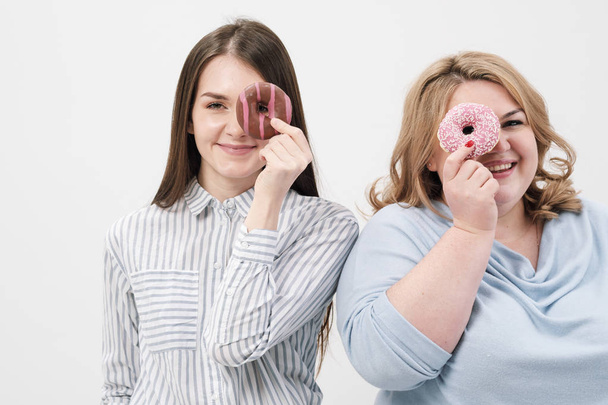 Two girls, thin and fat on a white background, are holding pink glazed donuts in their hands. - Foto, Bild