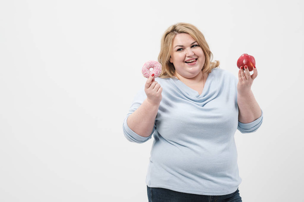 A fat, plump blonde woman on a white background, holding pink glazed donuts and a red apple. - Fotografie, Obrázek