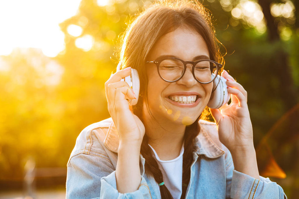 Pleased happy young teenage girl student sitting outdoors in beautiful green park listening music with headphones. - 写真・画像
