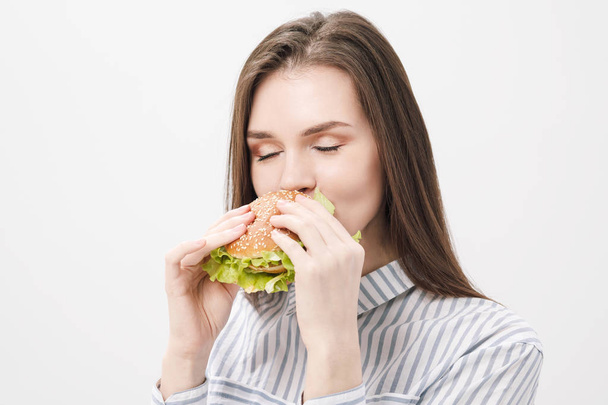 Young beautiful brunette girl on a white background with a hamburger in her hands. - Zdjęcie, obraz