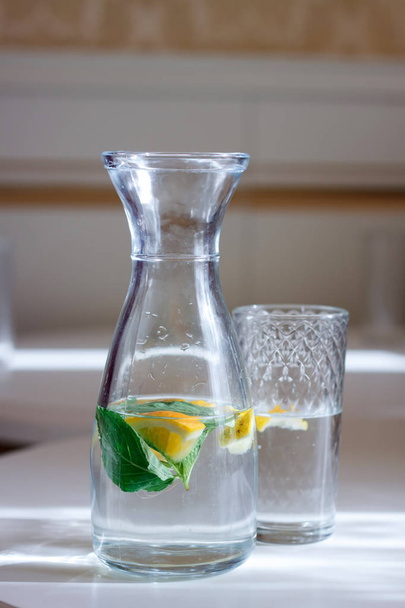 Крупный план бутылки с колпачком и стаканом воды с мятой и лимоном белый стол и солнечный свет, избирательный фокус
 - Фото, изображение