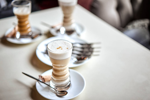 Latte glass with layered latte, cappuccino or mocha with foam on table in cafe with milk saucer and spoon. Three cups of coffee. Food and drink toning image. - Fotografie, Obrázek