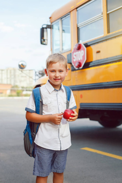 Divertido lindo niño estudiante caucásico feliz con mochila y manzana cerca de autobús amarillo el 1 de septiembre día. Educación y vuelta al concepto escolar. Alumno infantil listo para aprender y estudiar
.  - Foto, imagen