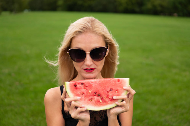 The girl falls in love on the field in photochromic glasses in the hand holding a slice of watermelon black hat from behind a beautiful forest. - Valokuva, kuva