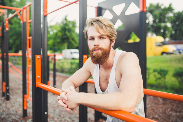Portre genç sakallı adam kamu spor zemin üzerinde duran ve paralel barlar üzerinde eğitim. Yakışıklı egzersiz atleti. Sakallı portre sportif adam, eğitimden sonra dinlenme. Yaz egzersiz açık havada - Fotoğraf, Görsel