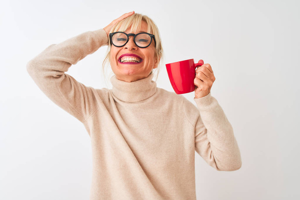 Mujer de mediana edad con gafas que beben taza roja de café sobre fondo blanco aislado estresado con la mano en la cabeza, conmocionado por la vergüenza y la cara sorpresa, enojado y frustrado. Miedo y malestar por error
. - Foto, imagen