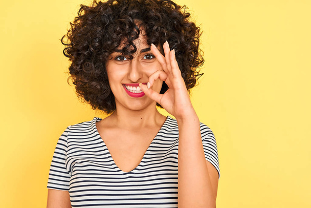 Nuori arabi nainen kihara tukka yllään raidallinen mekko yli eristetty keltainen tausta tekee ok merkki sormilla, erinomainen symboli
 - Valokuva, kuva