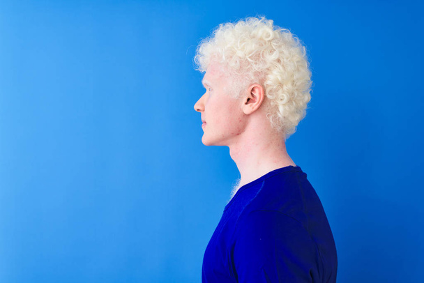 Mladý albín plavovlasý muž, který nosí neformální tričko stojící nad izolovaným modrým pozadím s pohledem na stranu, odpočívá v profilu s přirozenou tváří s sebevědomým úsměvem. - Fotografie, Obrázek