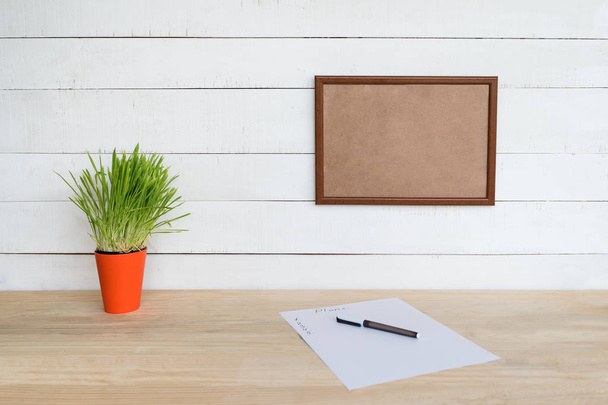 Empty frame on white wall. Sheet of paper and a pen, green houseplant on the table. Working space at home - Photo, Image