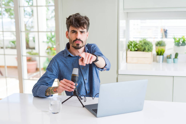 Young man recording podcast using microphone and laptop pointing with finger to the camera and to you, hand sign, positive and confident gesture from the front - Zdjęcie, obraz