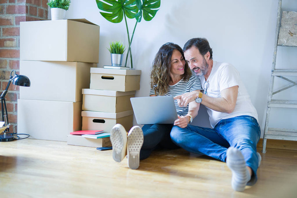 Orta yaş üst düzey romantik çift aşk etrafında kutuları ile daire katında oturan ve bilgisayar laptop kullanarak yeni bir eve hareket için mutlu gülümseyen - Fotoğraf, Görsel