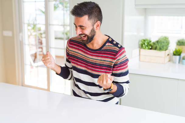 schöner hispanischer Mann, der zu Hause einen gestreiften Pullover trägt, sehr glücklich und aufgeregt Sieger-Geste mit erhobenen Armen macht, lächelt und schreit nach Erfolg. Festkonzept. - Foto, Bild