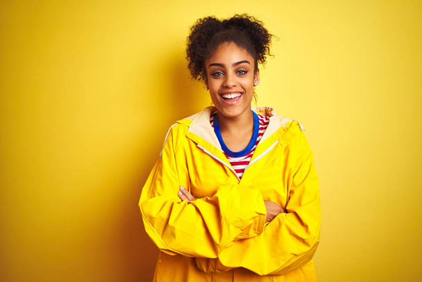 Young african american woman wearing rain coat over isolated yellow background happy face smiling with crossed arms looking at the camera. Positive person. - Φωτογραφία, εικόνα
