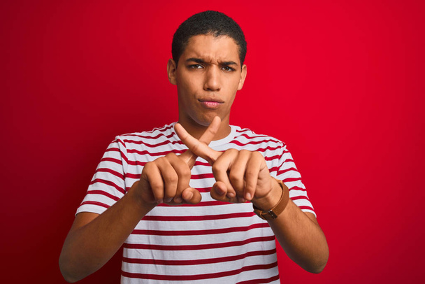 Νέος όμορφος Άραβας άντρας φορώντας ριγέ t-shirt πάνω από απομονωμένο κόκκινο φόντο απόρριψη έκφραση διασχίζοντας τα δάχτυλα κάνει αρνητικό σημάδι - Φωτογραφία, εικόνα