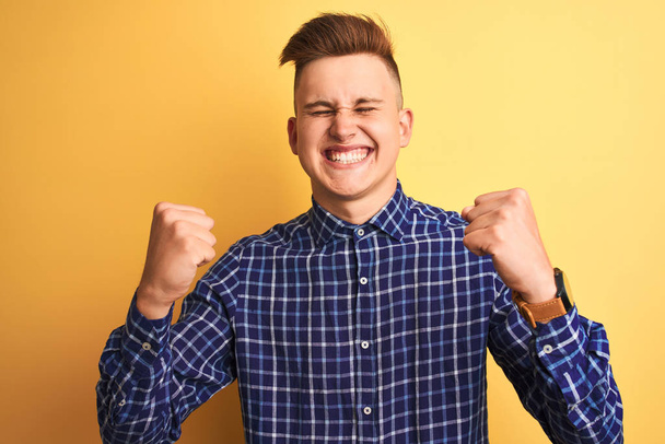 Young handsome man wearing casual shirt standing over isolated yellow background very happy and excited doing winner gesture with arms raised, smiling and screaming for success. Celebration concept. - Photo, Image