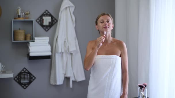 Face massage. Beautiful woman wrapped in towel after shower is getting massage face using jade roller massager - Filmati, video