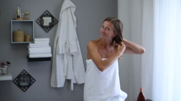 ヘアケアのコンセプト。シャワー後にタオルに包まれた美しい女性は、彼女の濡れた髪にココナッツオイルを適用しています - 映像、動画
