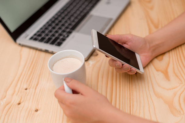 Young lady holding a smartphone and mug of tea. Gray laptop computer on the background with black keyboard a cup of coffee. Office supplies, technological devices and wooden desk. - Photo, Image