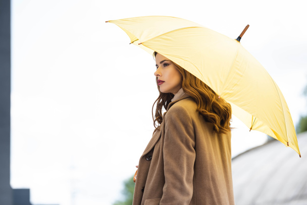 atractiva mujer en abrigo mirando hacia otro lado y sosteniendo paraguas amarillo
  - Foto, imagen