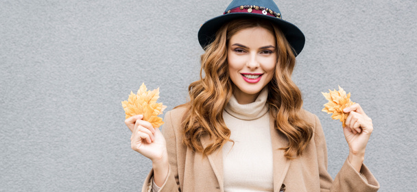 panoramic shot of attractive woman in blue hat looking at camera and holding yellow leaves  - Photo, image