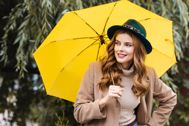 mavi şapkalı çekici kadın uzağa bakıyor ve sarı şemsiye tutuyor  - Fotoğraf, Görsel