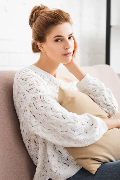 atraktivní žena v bílém svetru držící polštář a pohled na kameru  - Fotografie, Obrázek