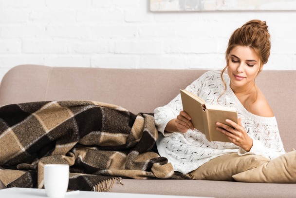 atraktivní žena v bílém svetru, který sedí na pohovce a čte si knihu  - Fotografie, Obrázek