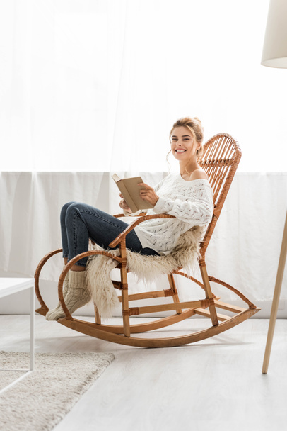 ελκυστική γυναίκα με λευκό πουλόβερ κάθεται σε κουνιστή καρέκλα και κρατώντας το βιβλίο - Φωτογραφία, εικόνα
