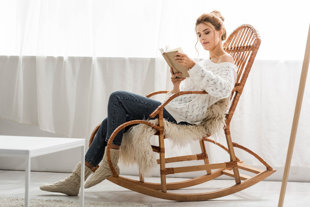 vonzó nő fehér pulóvert ül hintaszékben és olvasás könyv - Fotó, kép