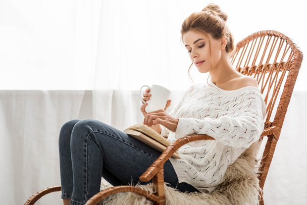 приваблива жінка в білому светрі, сидить на кріслі-качалці і читає книгу
 - Фото, зображення