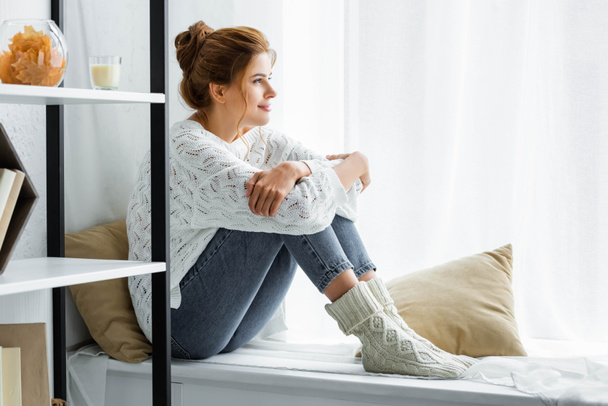 Seitenansicht einer attraktiven Frau in weißem Pullover und Jeans, die sitzt und wegschaut  - Foto, Bild