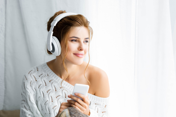 attraktive Frau im weißen Pullover, Musik hörend und Smartphone in der Hand  - Foto, Bild