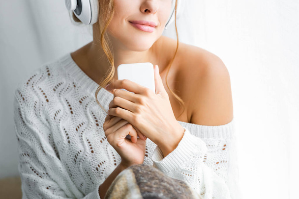 cropped view of woman in white sweater listening music and holding smartphone  - Photo, Image