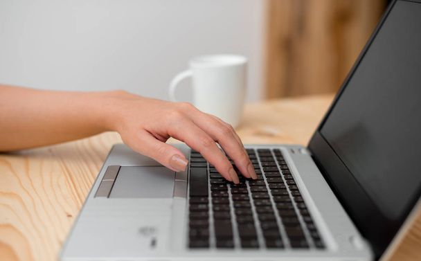 Mladá dáma používá šedý přenosný počítač a v pozadí si zadáváním černé klávesnice s šálkem kávy nebo čaje. Kancelářské potřeby, technologická zařízení a dřevěné psací stůl. - Fotografie, Obrázek