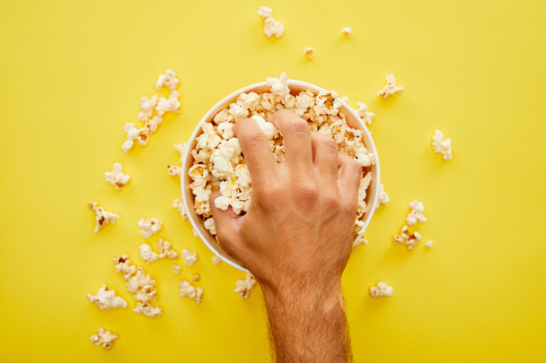 cropped view of man grabbing delicious popcorn from bucket on yellow background - Foto, imagen