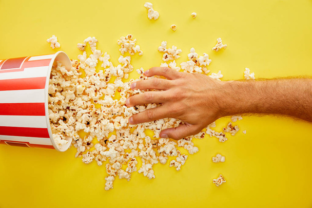 cropped view of man putting hand on delicious popcorn on yellow background - Фото, изображение