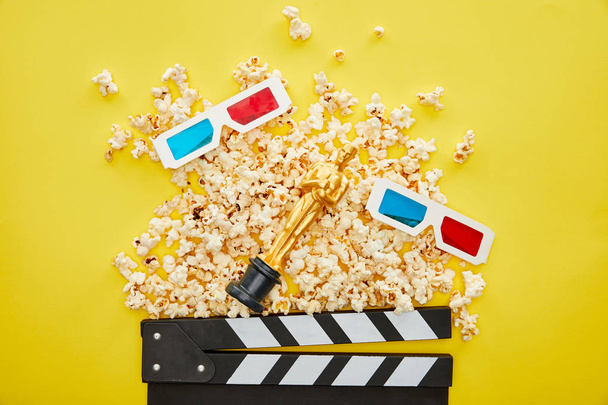KYIV, UKRAINE - AUGUST 13, 2019: top view of delicious popcorn, 3d glasses and clapper board with golden Oscar statuette on yellow background - Photo, image