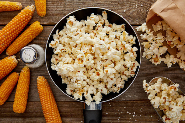top view of salty popcorn in frying pan near corn on wooden background - Fotoğraf, Görsel