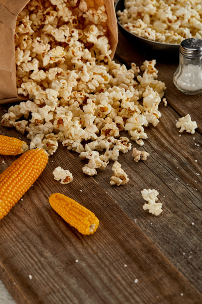 delicious popcorn scattered from paper bag near corn and frying pan on wooden background - Photo, Image